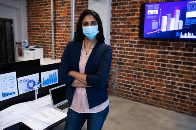 Portrait of biracial woman working in a casual office, wearing face mask, looking at camera and crossing arms. Social distancing in the workplace during Coronavirus Covid 19 pandemic.