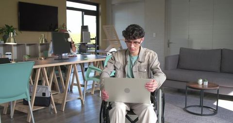 Young man in wheelchair working on laptop in modern coworking space promoting accessibility
