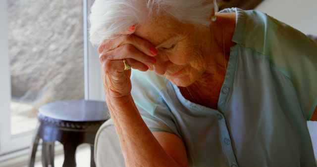 Elderly woman appears deep in thought while resting her head on her hand. The scene takes place indoors with natural light streaming through a window. This image can be used for themes related to aging, emotional health, senior living, and personal reflection.