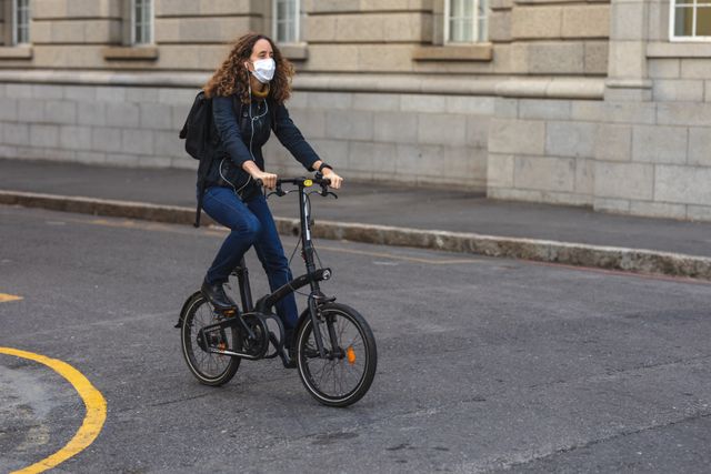 Caucasian woman out and about in the city streets during the day, wearing a face mask against coronavirus, covid 19, riding on bicycle building behind her.