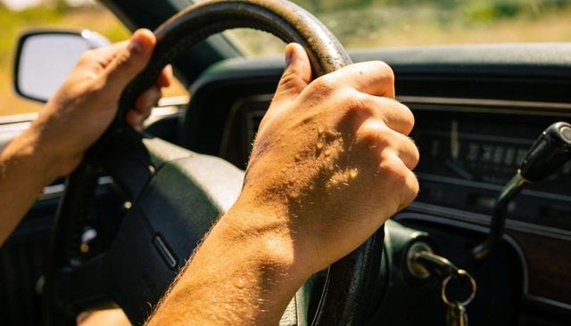 Man gripping worn steering wheel driving on sunlit rural road with keys in ignition