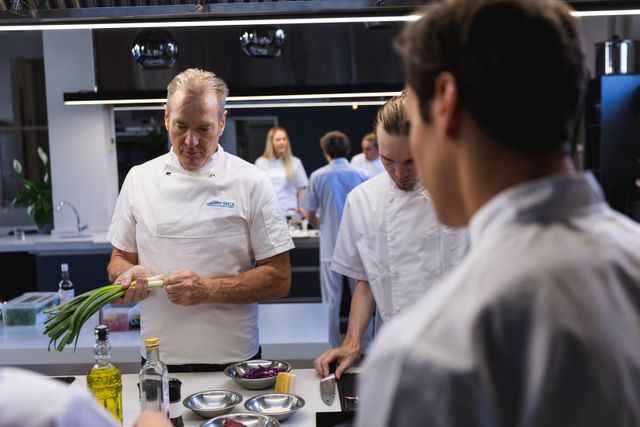 Group of Caucasian chefs cooking in a modern busy kitchen, male chef holding spring onions and instructing students. Cookery class at a restaurant kitchen. Workshop cooking food.