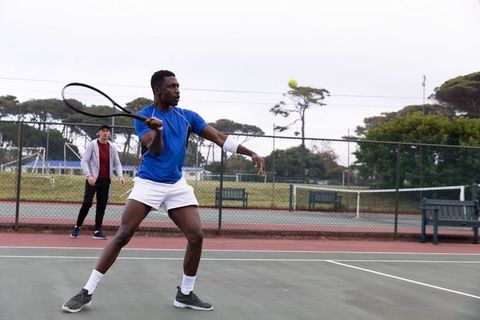 Black tennis player swinging forehand on outdoor hard court with partner observing during practice