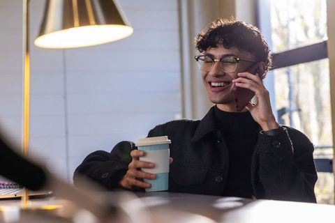 Young asian man smiling while talking on phone and holding coffee in cozy daylight cafe