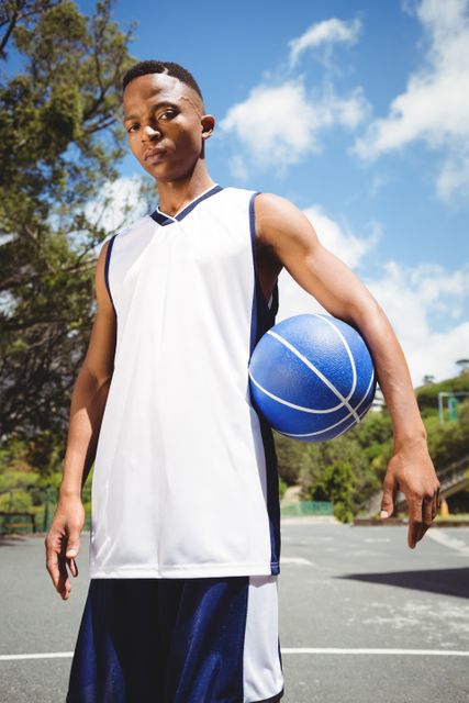 Teenage basketball player standing on an outdoor court, holding a basketball under one arm. Ideal for use in sports-related content, youth athletic programs, fitness promotions, and advertisements for sportswear or equipment.