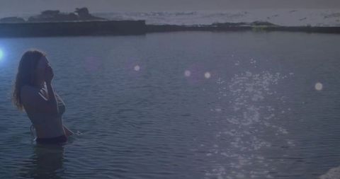 Backlit woman wading at sunrise touching face silhouette with sparkling water reflections