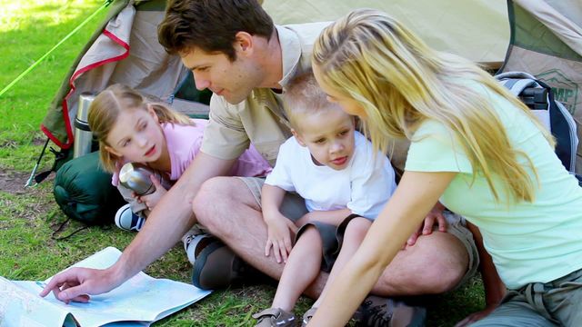 A family enjoys a day outdoors by examining a map together beside their tent. This heartwarming depiction of a family with young children highlights the essence of family bonding through the joys of camping. Extensive greenery surrounds the happy group as they plan their exploration, contributing to the outdoor adventure theme. Ideal for use in advertisements for camping gear, family travel experiences, or adventure outings aimed at capturing family involvement.