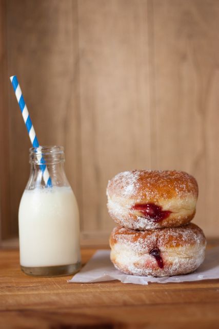Two sugar-coated jelly donuts on a parchment paper beside a small bottle of fresh milk with a blue striped straw. Perfect depiction of a delicious breakfast or sweet snack. Ideal for food blogs, bakery advertisements, and breakfast menus. The rustic wooden background adds a warm, homely feel.