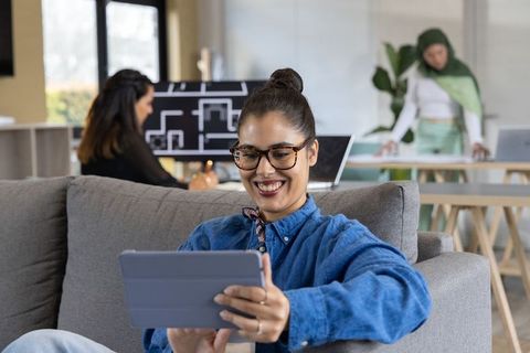 Smiling woman using tablet on sofa in modern coworking space with diverse team