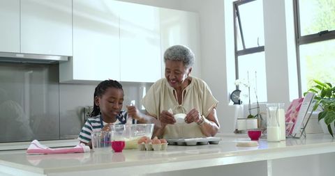 Grandmother teaching granddaughter baking and mixing batter at bright modern kitchen island