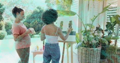 Young woman painting on sunlit porch with friend watching and holding mug garden terrace