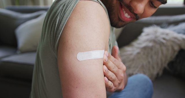 Man sitting on couch, displaying band-aid on upper arm, indicating recent vaccination. Can be used for vaccination campaigns, health promotion, preventive care advertisements, or content encouraging vaccines.