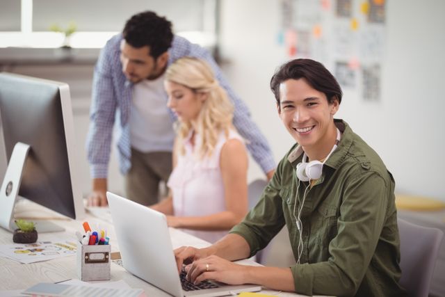 Young professional smiling while working on laptop in a creative office environment. Ideal for use in business, technology, and teamwork-related content. Perfect for illustrating modern workspaces, productivity, and collaborative efforts in a diverse team setting.