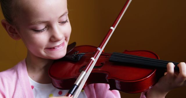 Young girl happily practicing violin with a focused yet joyful expression. Ideal for educational materials, music lesson ads, and promotional content for learning musical instruments. Can also be used in content related to children's hobbies, childhood development, and highlighting the importance of music in early education.