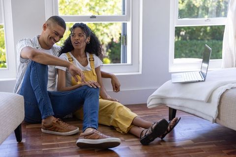 Young couple relaxing on hardwood floor in sunlit bedroom with laptop and conversation