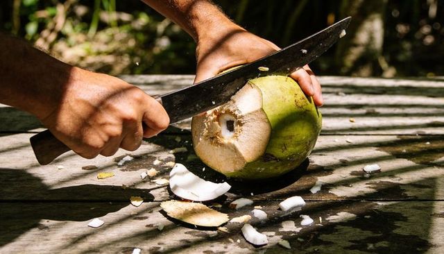 Hands cutting green coconut with machete on sunlit wooden table, tropical drink prep