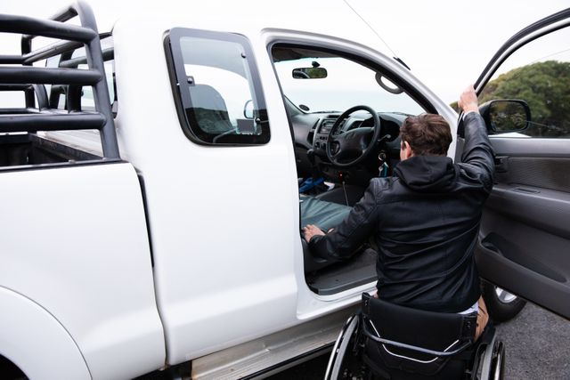 Rear view of a young Caucasian disabled man in a wheelchair beside his pick-up truck, parked with the door open, about to climb into the truck 
