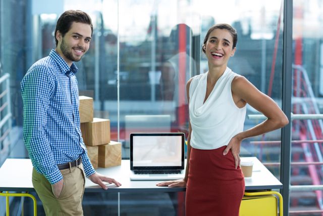 Two business executives, a man and a woman, are standing in a modern office environment. They are smiling and appear confident, suggesting a positive and collaborative work atmosphere. The man is wearing a blue checkered shirt and beige pants, while the woman is dressed in a white blouse and red skirt. A laptop and some boxes are on the table behind them. This image can be used for corporate websites, business presentations, team-building materials, and professional networking profiles.