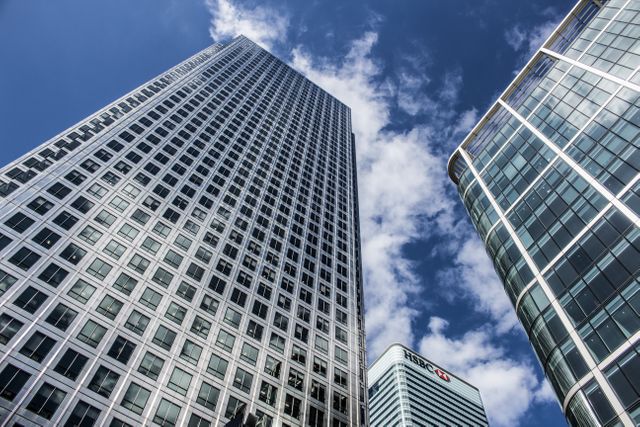 This image showcases modern office buildings in a bustling business district against a backdrop of a blue sky with clouds. The photo captures the elegance and grandeur of corporate skyscrapers and glass architecture, making it suitable for illustrating concepts related to business, finance, urban development, and architectural design. Great for use in marketing materials, real estate brochures, finance presentations, and corporate websites.