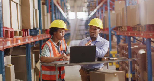 Two warehouse workers are discussing logistics while using a laptop amidst the aisles of a well-organized warehouse. Both are wearing safety gear, including yellow hard hats and an orange vest, emphasizing a safe working environment. This image can be used for materials related to warehouse management, logistics services, business operations, technology in supply chain, and teamwork in industrial settings.