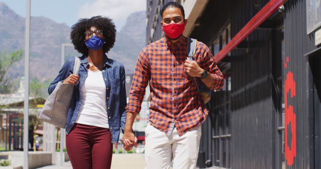 Diverse couple wearing face masks walking and holding hands. digital nomads on the go during coronavirus covid 19 pandemic.
