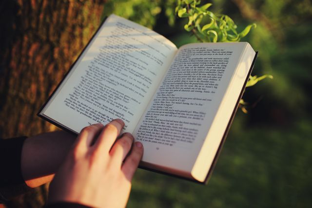 Photo depicts hands holding an open book while reading outside, with greenery in the background. Suitable for use in educational content, nature-related materials, or promoting reading and literacy. Ideal for blog posts about relaxing outdoor activities, digital magazines, or social media posts encouraging personal development and learning through reading.
