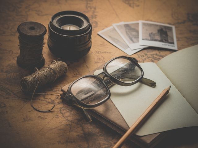 Items laid out on an antique map include round-rimmed glasses, a notebook and pencil, a camera lens, spools of thread, and vintage photographs. Suitable for travel blogs, adventure narratives, nostalgic themes, vintage photography collections, and creative writing materials.