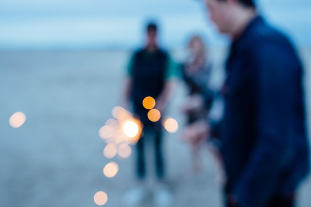 People holding sparklers by a beach, out-of-focus and creating a bokeh effect with the lights. Suitable for themes of celebration, friendship, beach events, festive activities, and relaxed summer evenings by the sea.