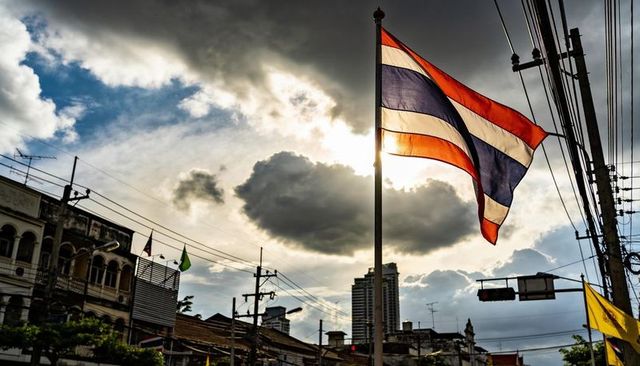 Backlit thai flag waving above bustling urban street with dramatic clouds and power lines