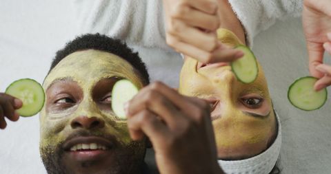 Diverse couple relaxed with face masks and cucumber slices at home spa