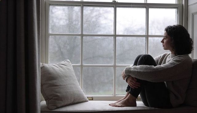 Woman sitting on window seat hugging knees looking out rainy window contemplative mood