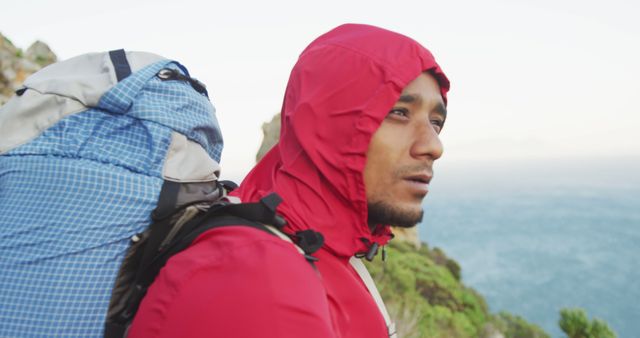 Young man wearing red jacket and carrying a blue backpack, hiking near mountains. Ideal for content on outdoor adventures, hiking gear, travel destinations, and active lifestyles. Can be used in travel blogs, adventure magazines, and advertisements for outdoor products.