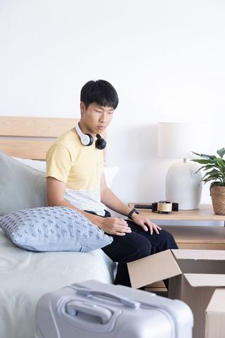 Asian man sitting on bed edge packing cardboard box and suitcase in minimalist modern bedroom