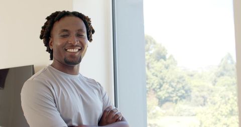 African american man smiling with crossed arms by sunlit floor-to-ceiling window interior