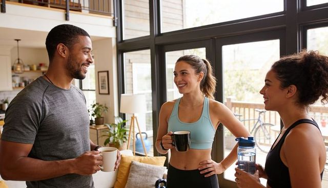 Smiling friends sharing coffee and water after home workout in sunlit living room