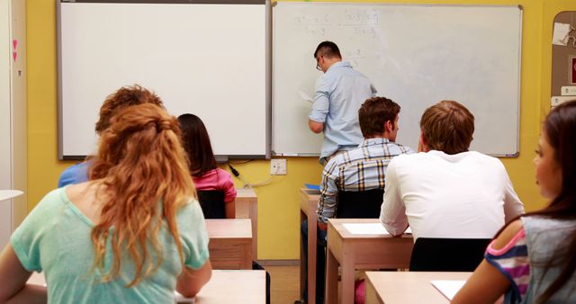 Teenage students are paying attention in their math class as the teacher writes on the whiteboard at the front of the room. The setting is a typical high school classroom with rows of desks and a whiteboard filled with equations. This subject is suitable for educational websites, school brochures promoting student involvement, and any content discussing secondary education and teaching methods.