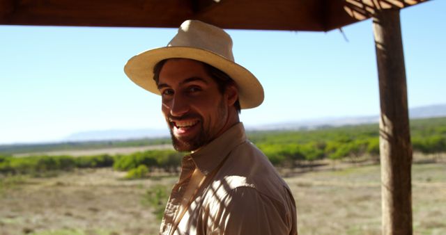 Man smiling while wearing a wide-brimmed hat in an open, natural landscape. Perfect for use in travel advertisements, adventure and outdoor lifestyle promotions, or leisure and tourism brochures depicting exploration and a connection with nature.