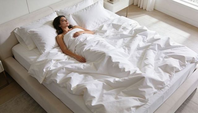 Woman reclining on white duvet in minimal sunlit bedroom, serene morning relaxation