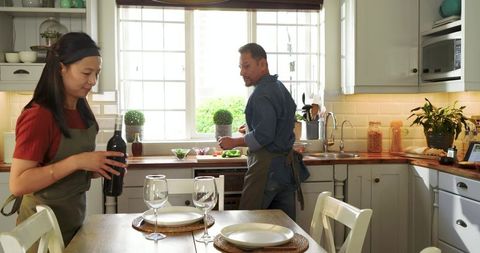 Asian couple preparing dinner in bright kitchen pouring wine and chopping vegetables