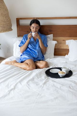 Woman in blue satin pajamas sipping coffee while sitting cross-legged on white bed