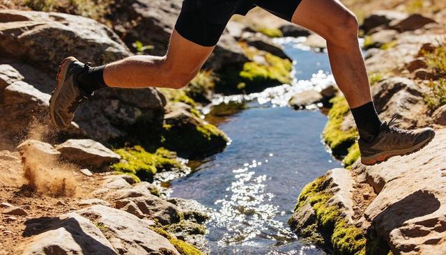 Leaping trail runner clearing mountain stream on rocky moss-covered path in sunlight