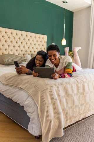Diverse women laughing on bed using tablet and phone in cozy bedroom with tufted headboard