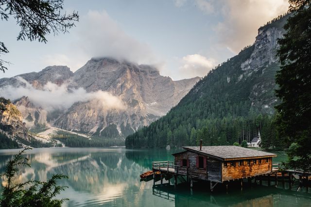 Serene scene of a wooden cabin perched at the edge of a calm lake, surrounded by misty mountains at sunrise. Reflection of mountains and cabin in the water enhances the tranquil beauty. Ideal for promoting travel destinations, outdoor adventures, and peaceful retreats.