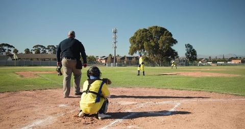 Youth baseball catcher squatting at home plate with umpire and infielders on sunny field