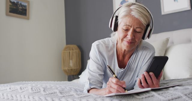 Senior woman lying on bed listening to music through headphones while writing in notebook. Ideal for depicting relaxation, senior lifestyle, enjoyment of hobbies, and personal moment of reflection. Suitable for wellness, leisure activity, and elderly care themes.