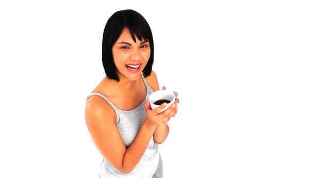 A young woman holds a coffee cup and smiles energetically against a plain white background. Ideal for use in marketing materials related to coffee products, energetic and positive lifestyle promotions, or advertising for cafes and breakfast businesses. The minimalist background provides ample space for copy or logos, making it versatile for various design needs.