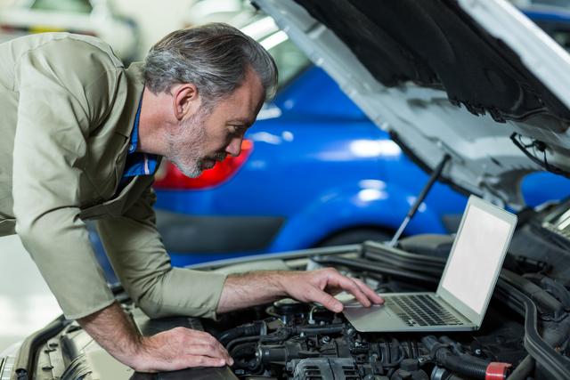 Mechanic using laptop while servicing car engine in repair garage