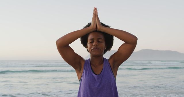 African woman practicing yoga in meditative pose on beach at sunrise, expression showing focus and calm. Ideal for wellness articles, meditation guides, outdoor fitness promotions, or content related to mental health and relaxation.