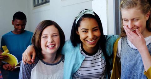 Teen friends laughing and linking arms in bright school hallway, multicultural group