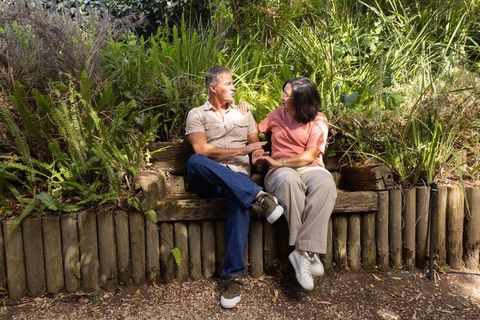 Diverse senior couple sitting on log bench holding hands in lush garden sharing conversation